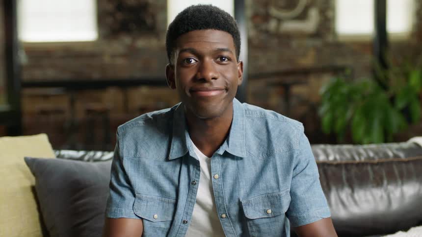 A young black man smiles into the camera while sitting on a sofa holding a game controller