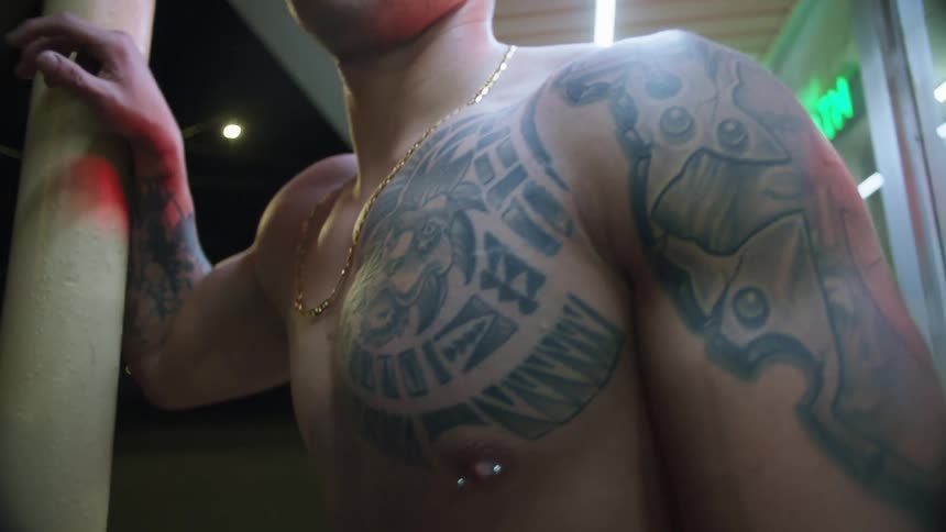 A shirtless tattooed man leans against a pole outside of a store at night