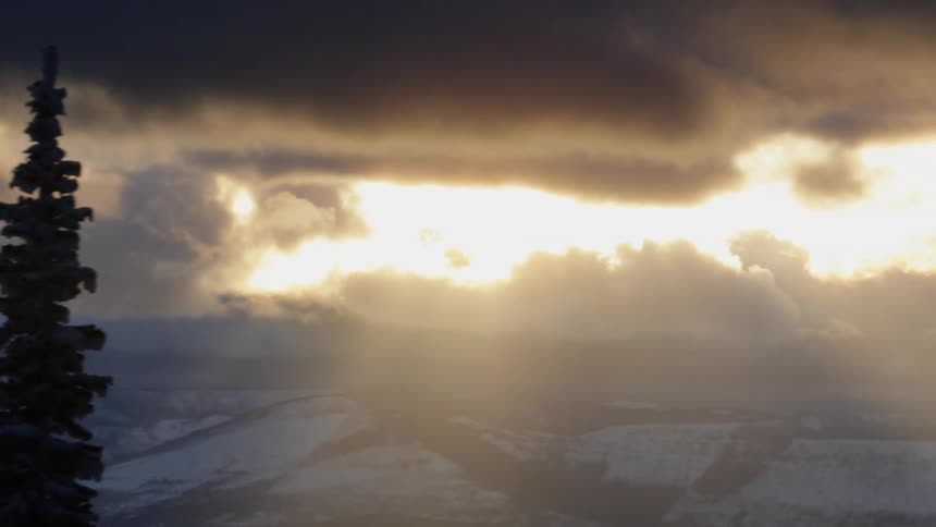 The sun shining through dark clouds over snow covered hills