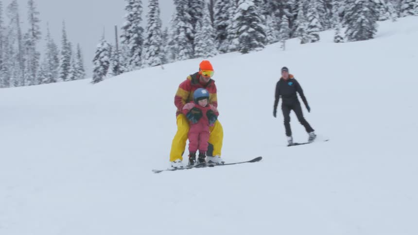 A father skis with his daughter down a snowy slope