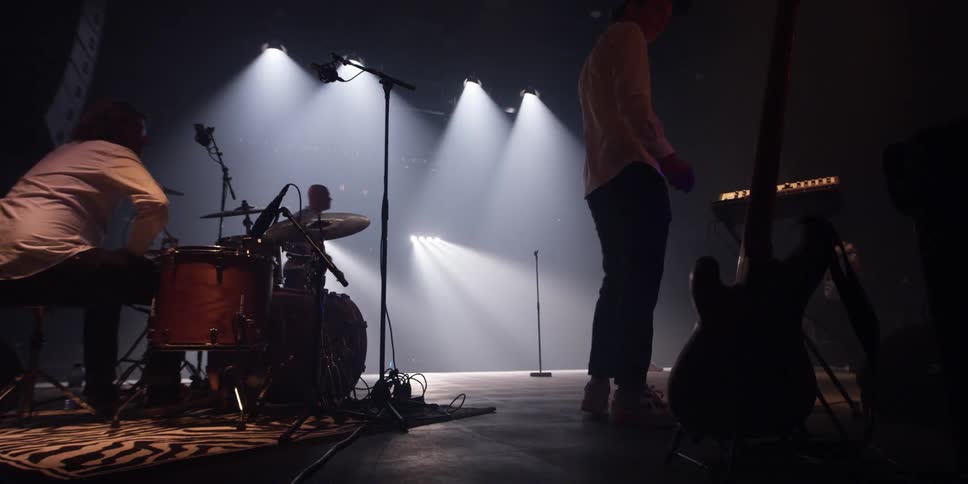Musicians hover near musical instruments as men walk across stage under spotlights