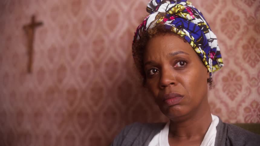 A woman wears a colorful headscarf as she sits in a wallpapered room with a hanging cross