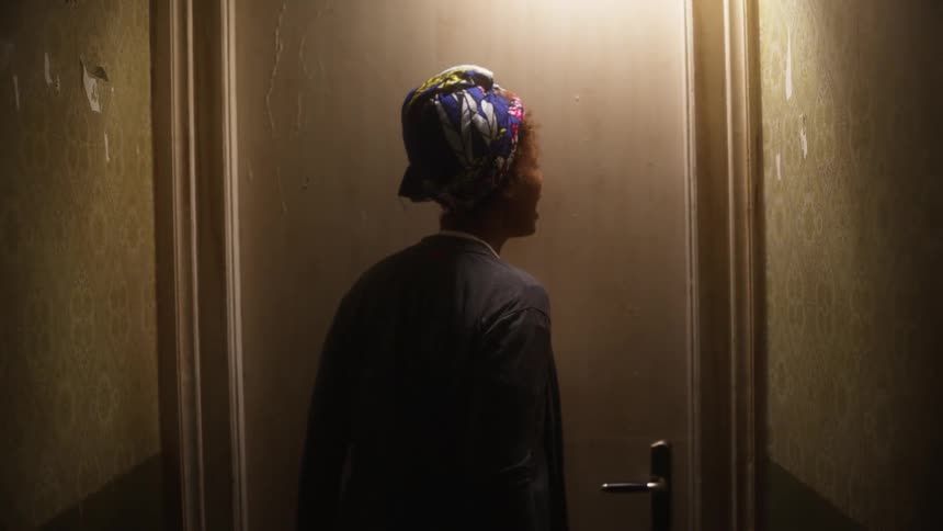 A woman wearing a headwrap angrily strikes and calls through a closed door