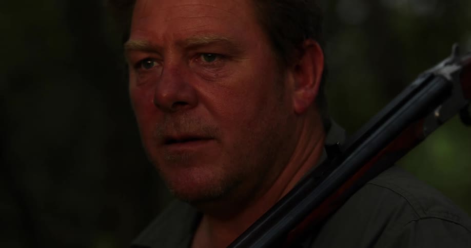 Close up of a mans face holding a shotgun on his shoulder in a forest at sunset