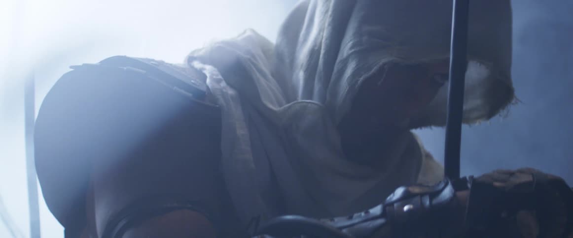 A hooded man looks down in a dark room