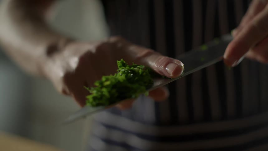 Man uses his finger to push freshly chopped herbs from the surface of a kitchen knife into a bowl
