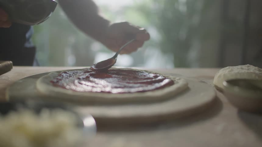 A person uses a spoon to spread sauce on a homemade dough