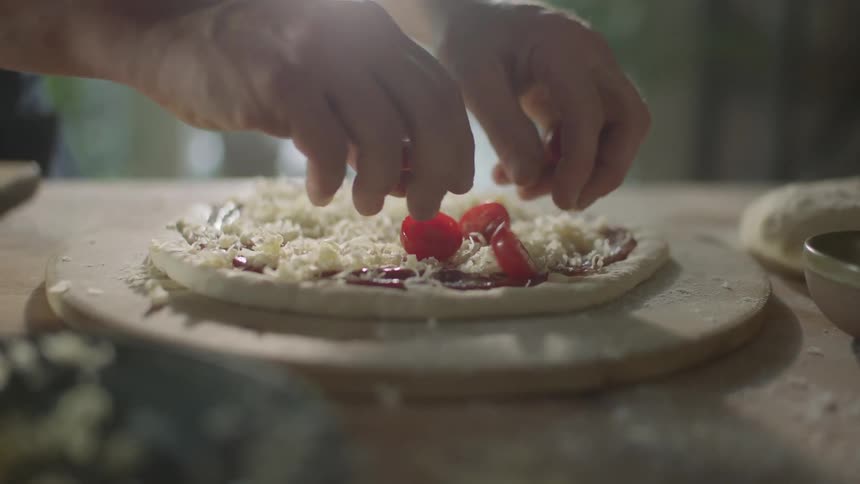 Male adds halved cherry tomatoes to the top of a homemade pizza that is sitting on top of a round wooded board dusted with flour