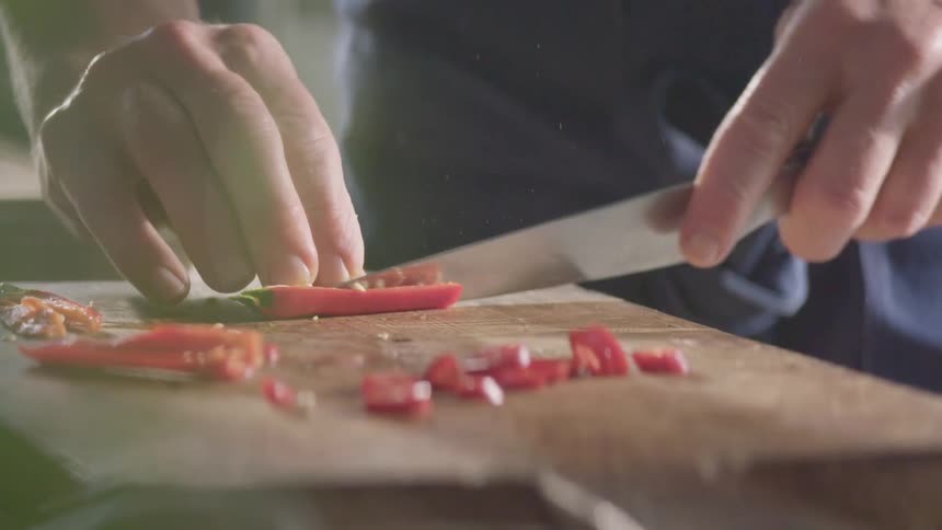 Cook with a black shirt cuts a red hot chili pepper with a knife on a cutting board in the kitchen