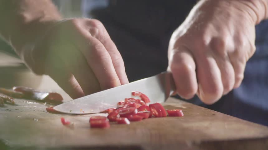 Cook with a black shirt cuts a red hot chili pepper with a knife on a cutting board in the kitchen