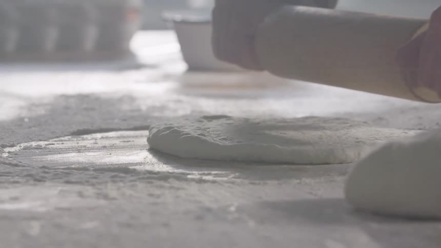 Person rolls pizza dough out of flour covered wooden surface using rolling pin
