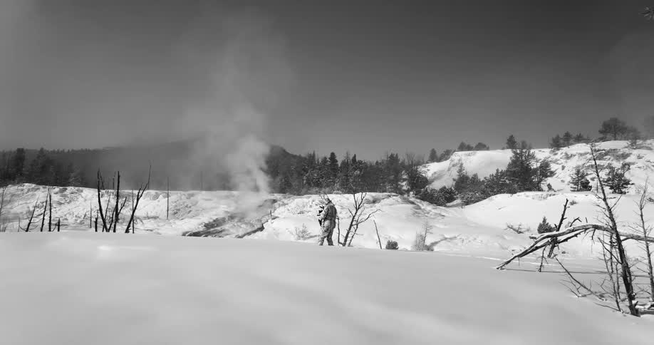 Man in full winter gear sets up a camera on a tripod in the snow on the hills