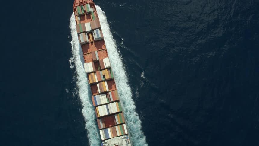 Freighter with deck stacked with shipping containers cuts through rippling water leaving wake