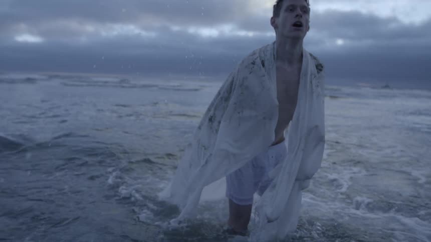 A man removing a white sheet from his head as he wades out of a rough ocean on a cloudy day