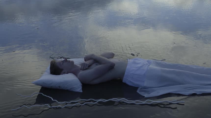 A white sheet is slowly pulled off a person lying on the wet sand