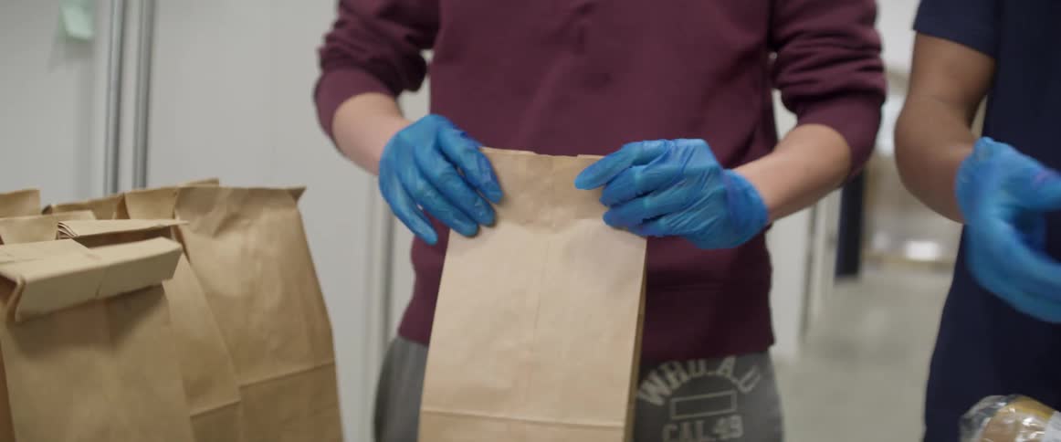 Two people place food inside brown paper bags on a table