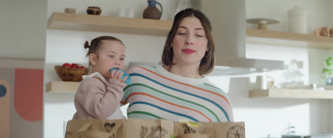 A woman holds her daughter as she tries to unload groceries from paper bags