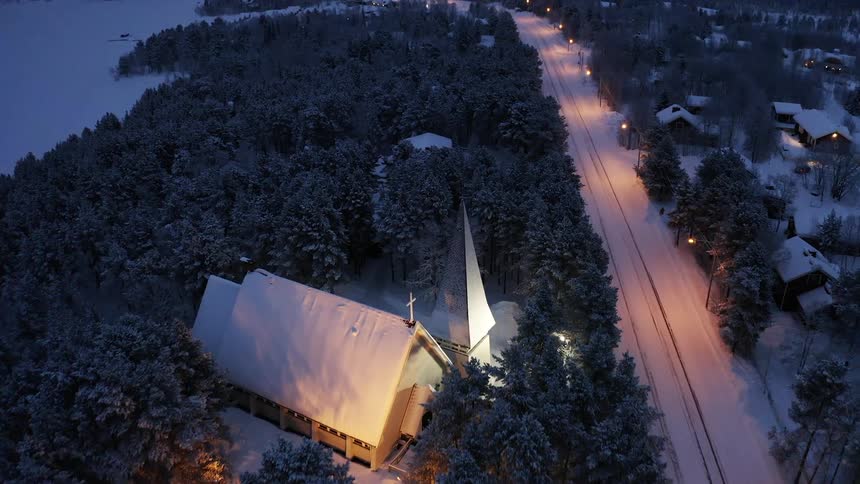 A church covered in snow and surrounded by trees next to a road with houses
