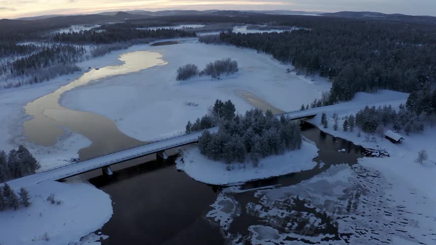 A bridge spans across a partially frozen river near the forest in winter as the day dawns