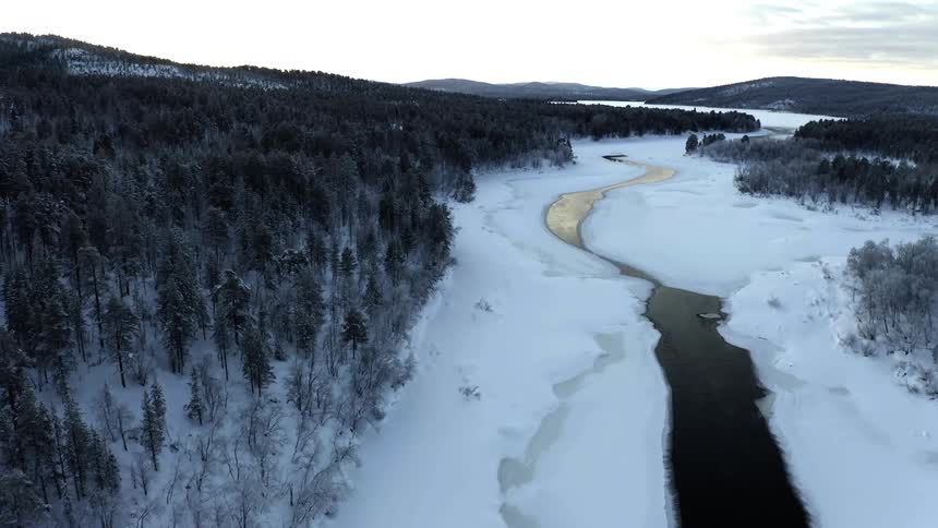 Aerial view of a nearly frozen river surrounded by snow covered pine forest at sunset