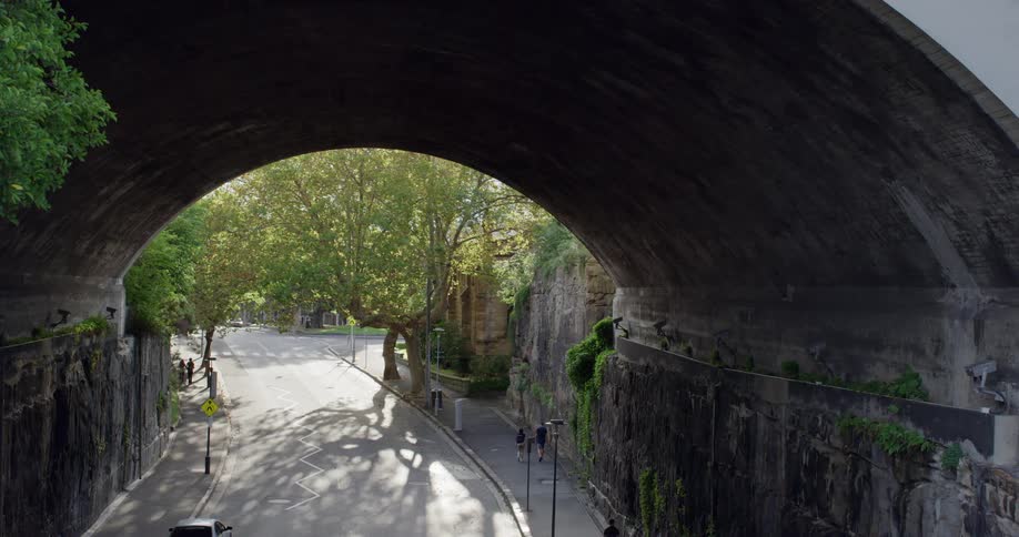 An arched bridge over a street with people walking on sidewalks and trees growing on a sunny day