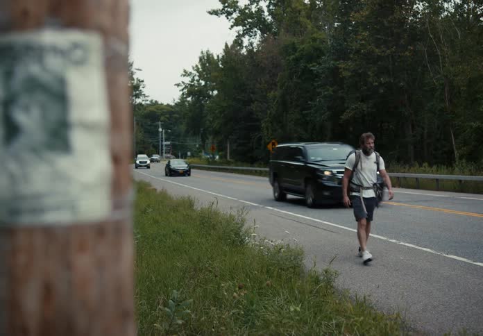Man with backpack walking on a rural road next to a missing person sign on a wooden pole