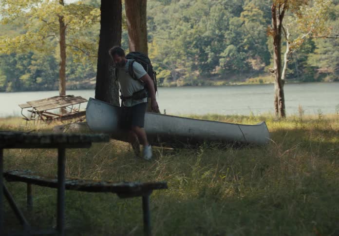 Bearded man with a backpack drags a kayak in the grass near a lake