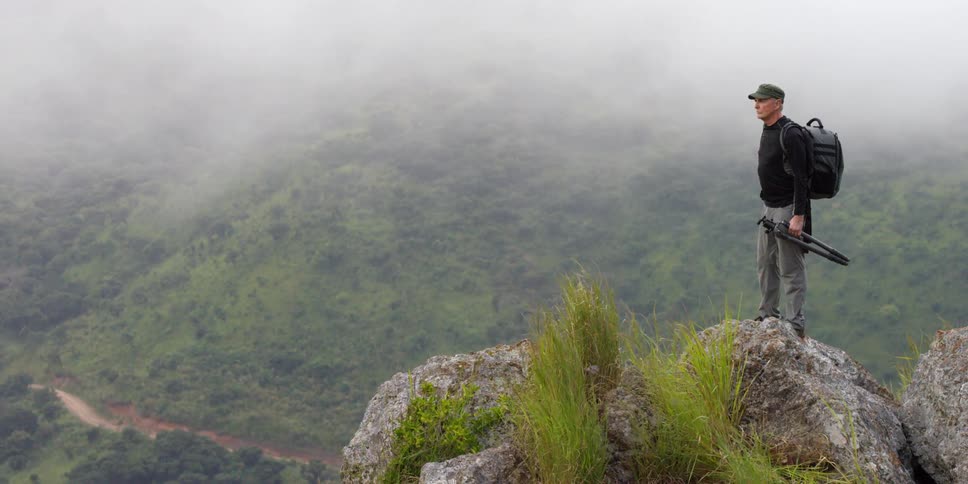 A man holding a tripod while standing on top of a boulder