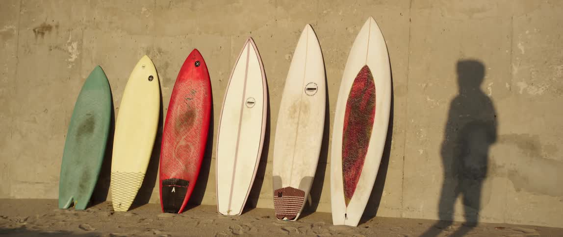 Six surfboards lean against a wall in the sand next to the shadow of a person