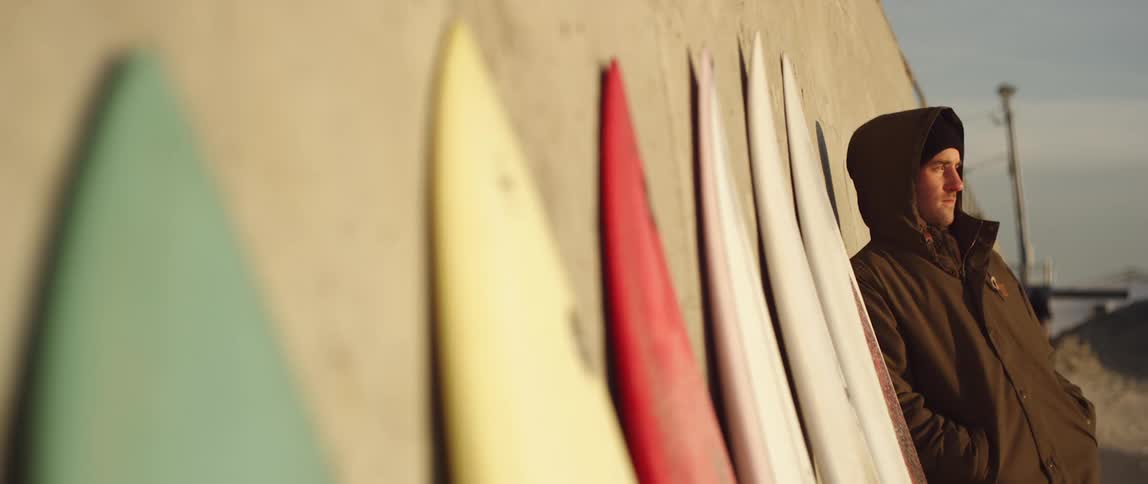 A white man in a coat leans against a wall next to a row of surfboards