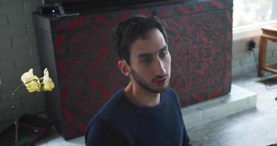 A middle eastern man sits on the floor in a living room and speaks