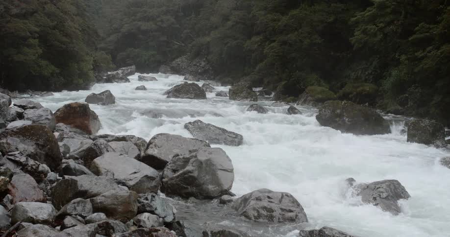 Rain falls on river rapids bounded by rocky vegetation dotted slopes