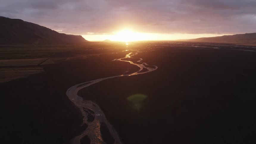 The low sun on the horizon shines beneath the cloudy sky to illuminate a winding stream through a barren field