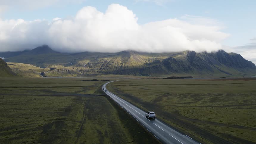 Vehicles travel along a curving road through a grassland towards a mountain range in the distance