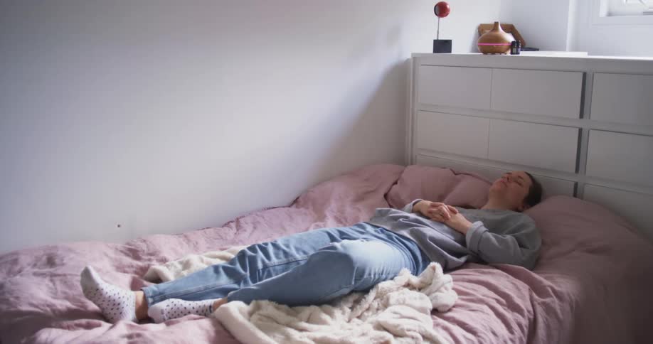 A woman lies on top of her covers on her bed napping fully clothed as her foot twitches