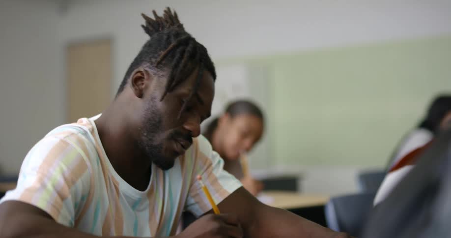 A male student sitting at a desk with a pencil writing on a notebook with a female student in the background