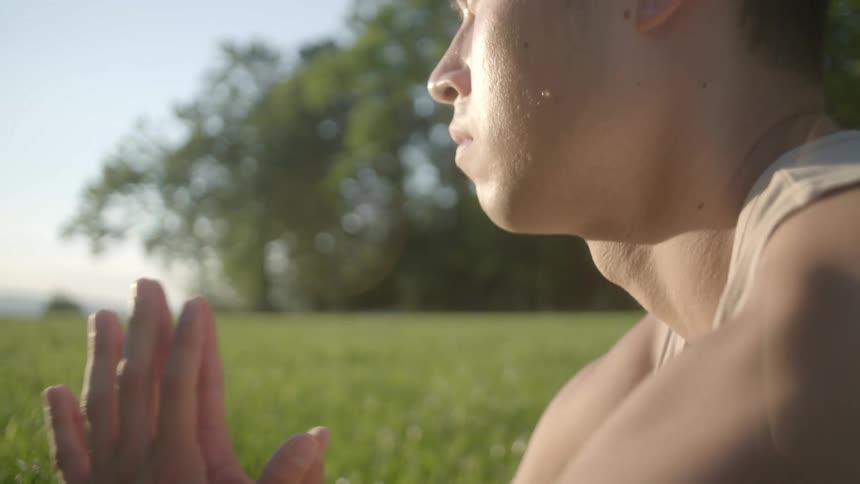A man sitting in the grass with hands pressed together bows his head on a bright sunny day