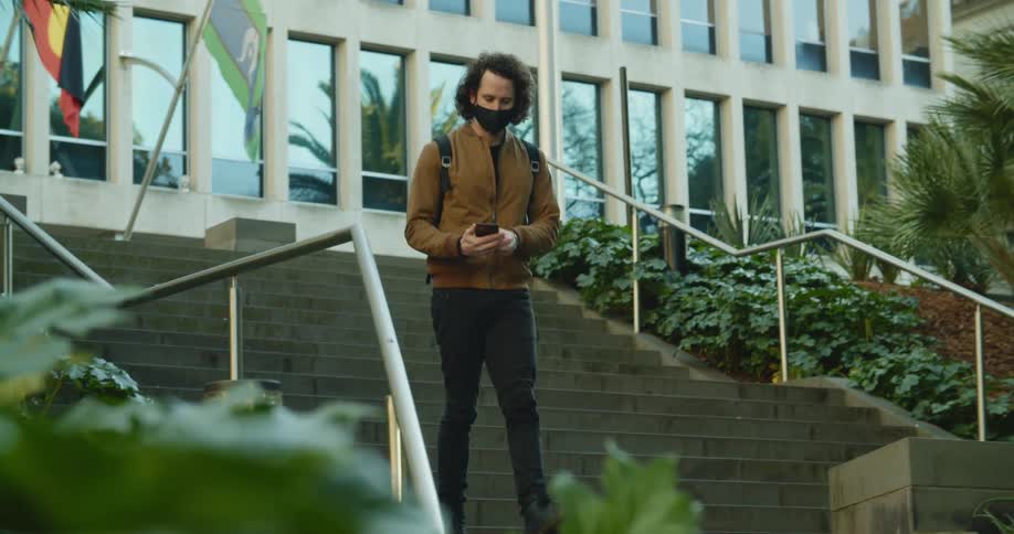 A man wearing a mask answers his phone while walking down the steps outside of a building