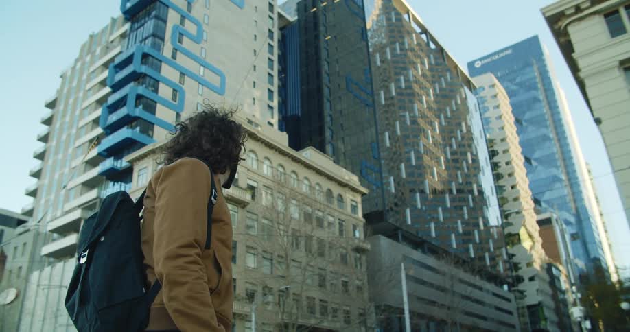 A man wearing a mask and a backpack stands on a city street looking up and around at buildings