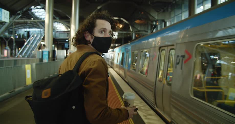 Man walks along transit station as overhead train with face mask wearing riders passes