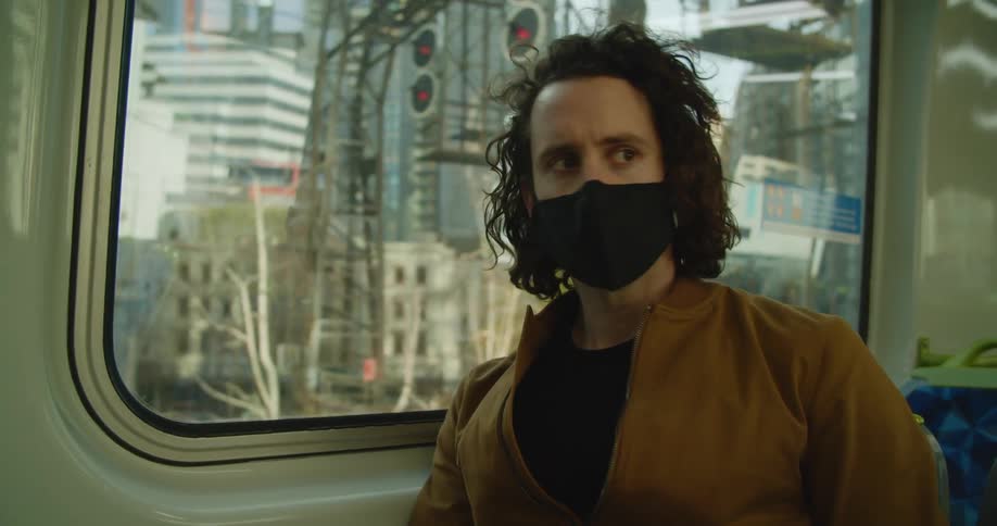 A man with a mask on a train next to a window looking at a smartphone