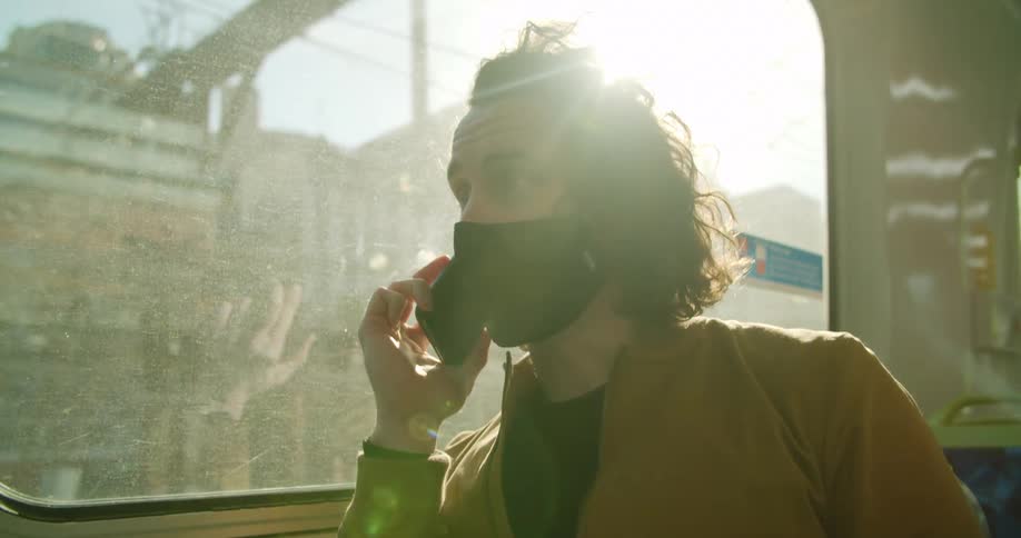 A man sits on a city bus talking on a cell phone and wearing a medical mask