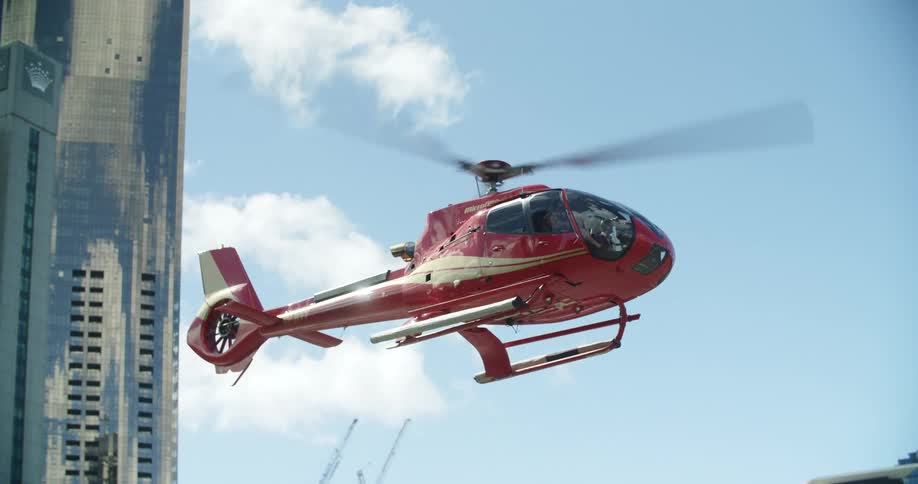 A red helicopter rises up with city buildings behind on a bright blue sky day