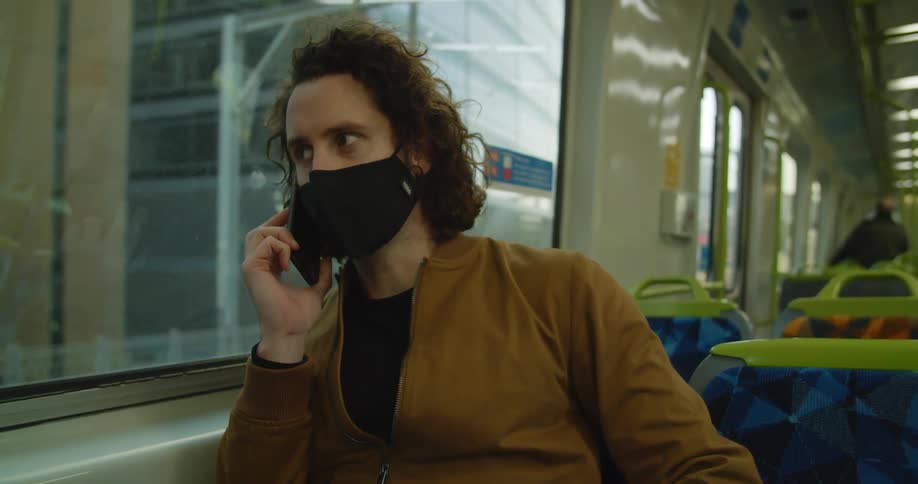 Man wearing face mask and jacket sits in public transit car talking on mobile phone passing buildings