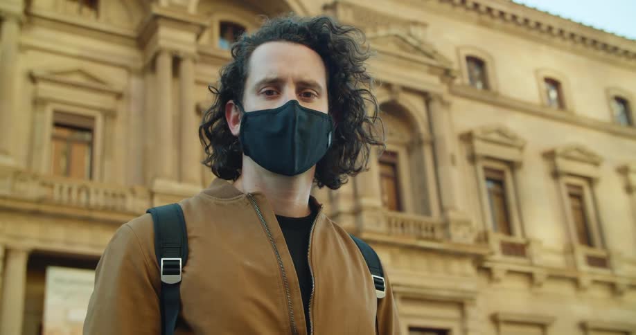 A teenager wearing a mask stands in front of a large building in a city