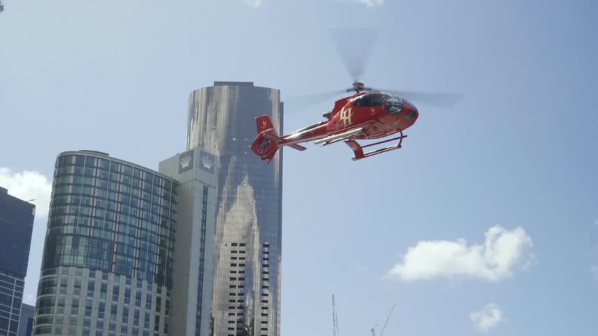 A red helicopter flies past and above tall skyscrapers in a city