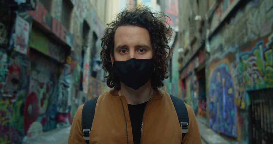 A man wearing a black face mask stands in a city alley filled with buildings marked with graffiti