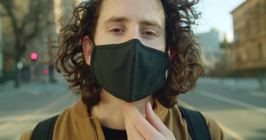 A man puts on a black medical mask and looks into the camera while standing in a city street