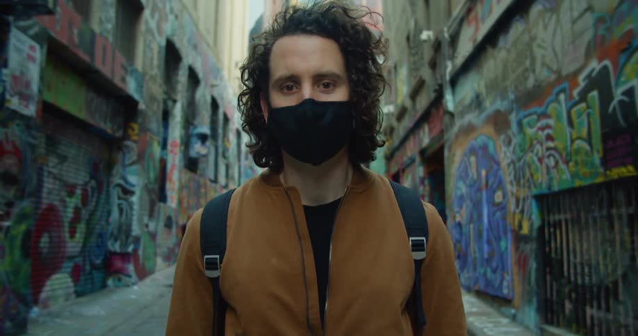 A young man wearing a mask stands in a street with buildings covered in graffiti