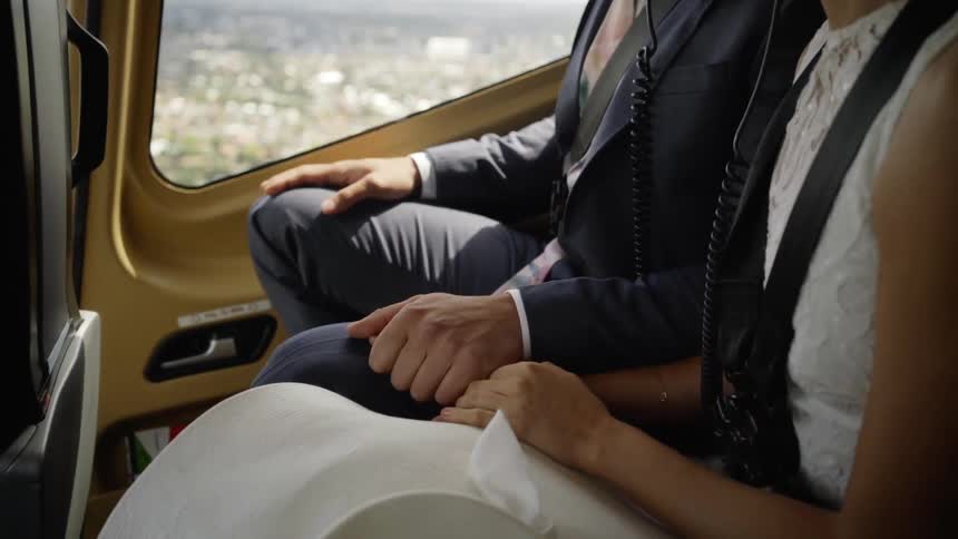 A man in a suit and a woman in a white dress hold hands while sitting in a helicopter flying over a city